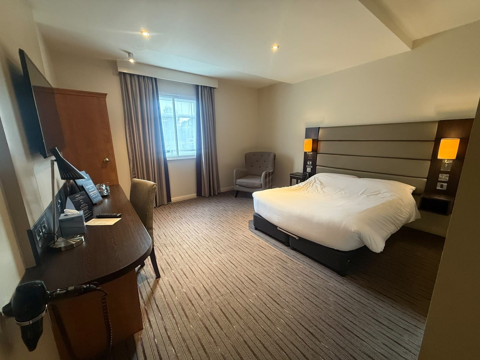 Habitación de hotel con una cama, un escritorio con teléfono, una silla, una ventana con cortinas y una lámpara de pared.