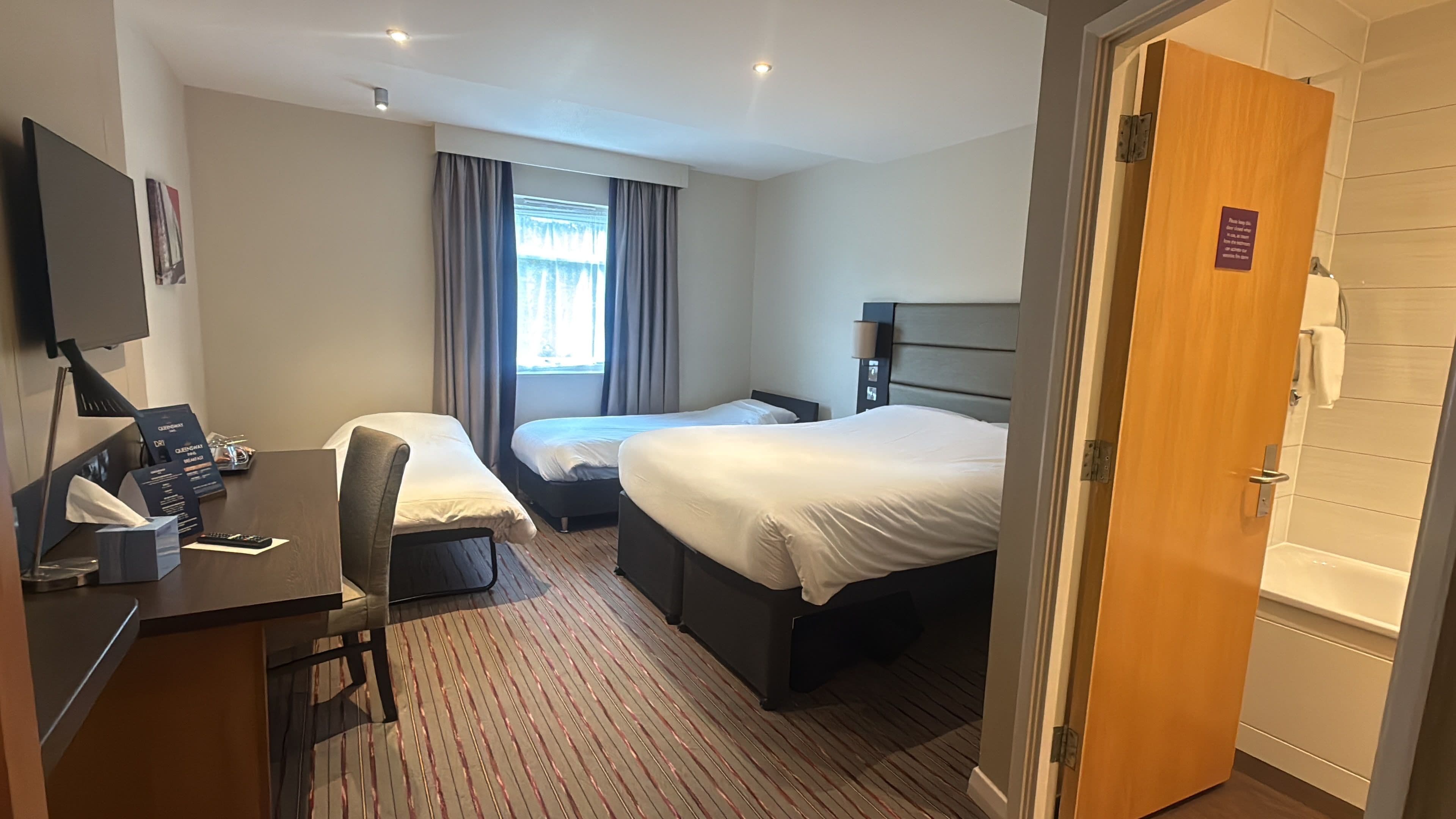 A hotel room with two beds, a desk with a computer, a TV, and a door leading to a bathroom.