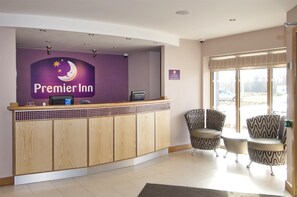Ein Hotel-Empfangsbereich mit einem 'Premier Inn' Schild, einem Empfangstresen, zwei gemusterten Sesseln und einem großen Fenster.