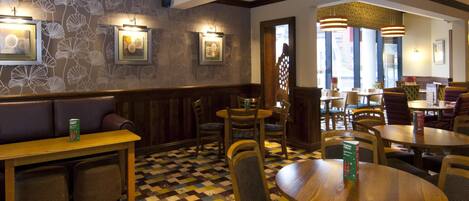 L’intérieur d’un restaurant est décoré de papier peint à motifs, de tables et de chaises en bois, et les murs sont ornés de tableaux encadrés.