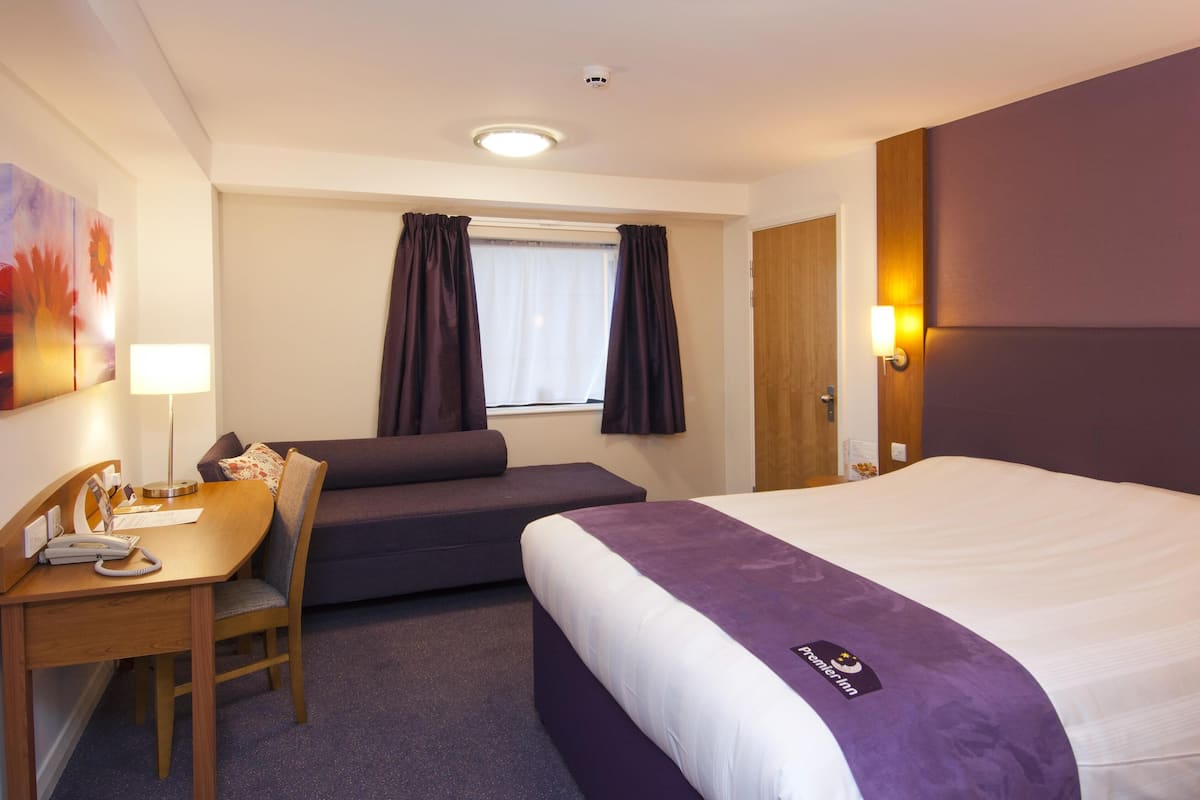 A hotel room with a bed, a desk with a telephone, a chair, a window with curtains, and a wall-mounted lamp.