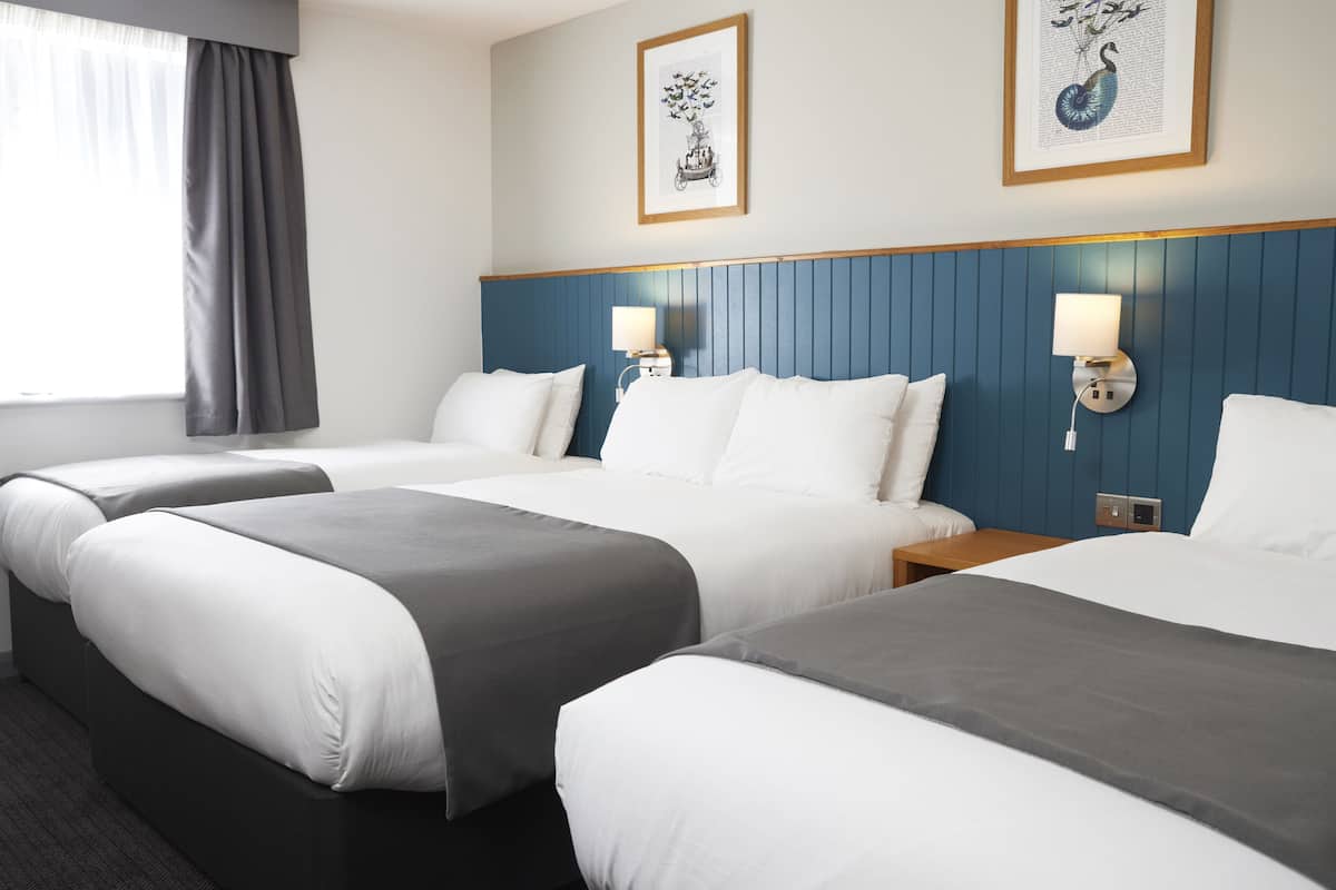 A hotel room with three beds, each with white and gray bedding, a blue headboard, and two wall-mounted lamps.