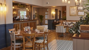 A restaurant with wooden tables and chairs, a bar area with glasses and bottles, and framed pictures on the walls. - Holmfield Arms By Greene King Inns (Wakefield)