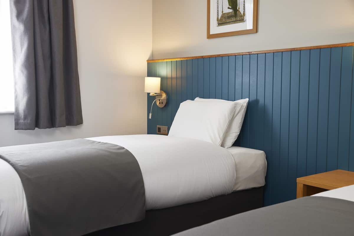 A hotel room with a bed, a nightstand, a wall-mounted lamp, and a window with curtains.
