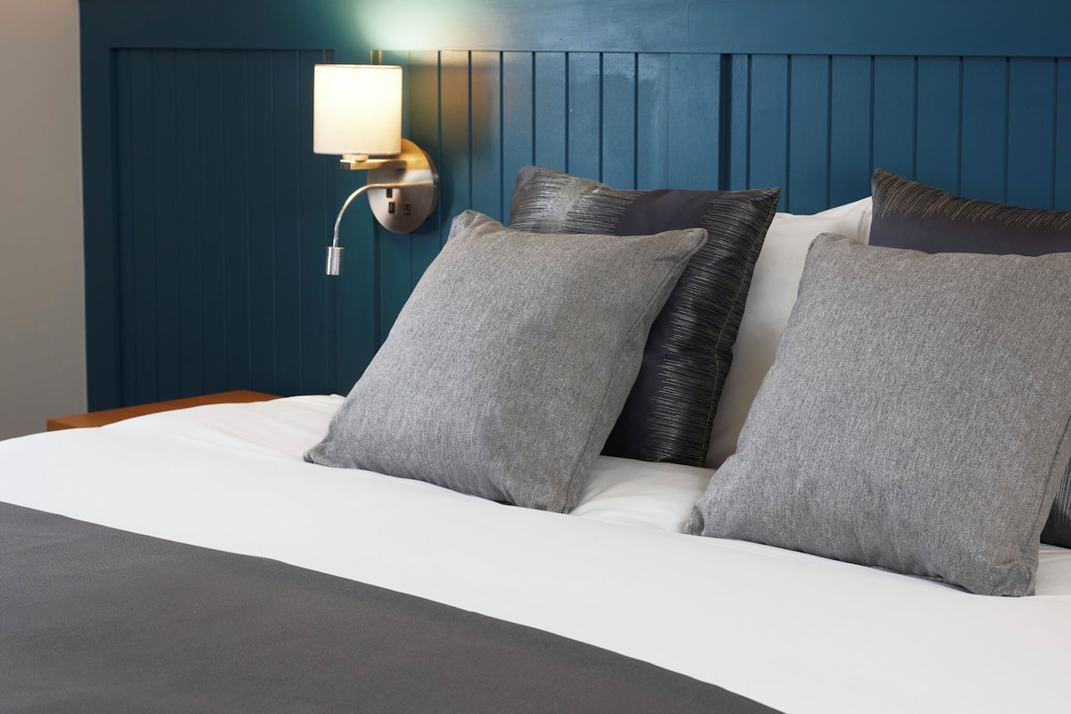 A hotel room with a bed, pillows, a wall-mounted lamp, and a framed picture above the bed.