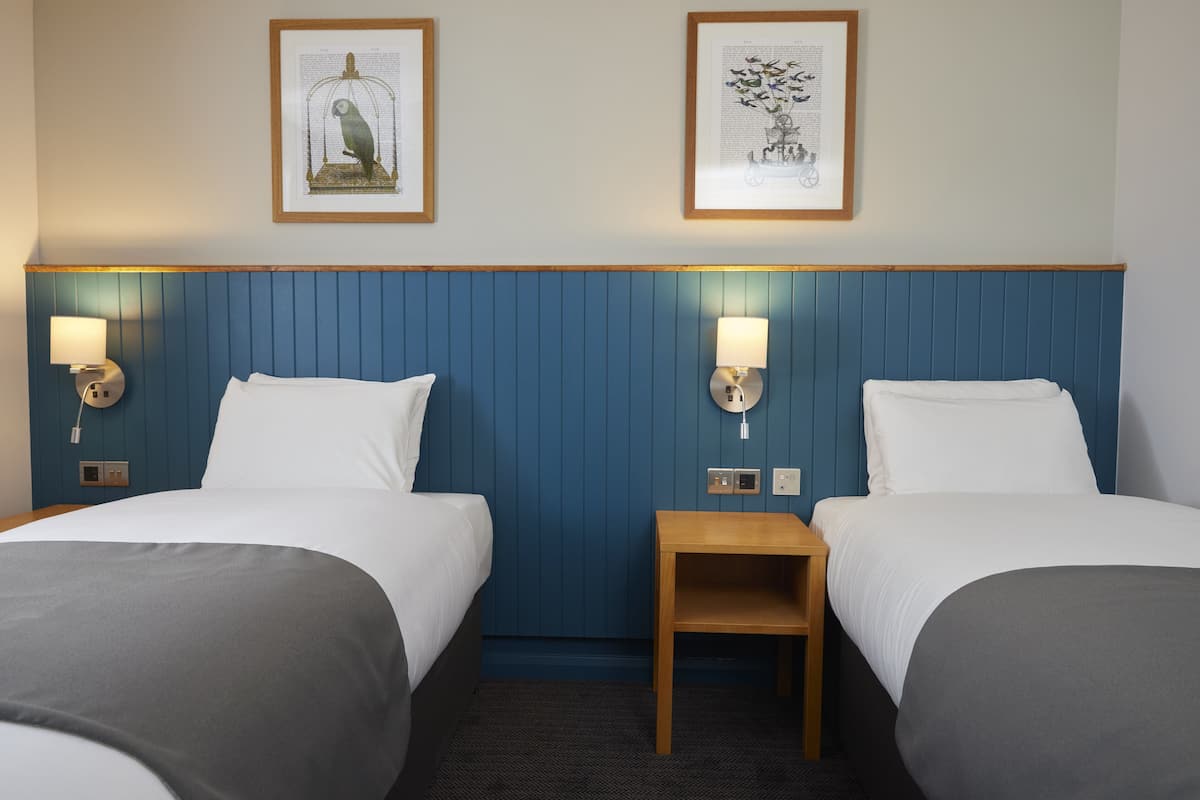 A hotel room with two beds, a wooden nightstand, wall-mounted lamps, and two framed pictures on the wall.