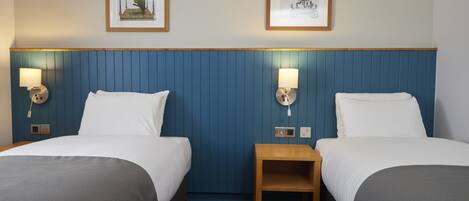 A hotel room with two beds, a wooden nightstand, wall-mounted lamps, and two framed pictures on the wall.
