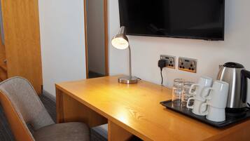 Habitación de hotel con escritorio de madera, televisor montado en la pared, lámpara, bandeja con vasos y tazas, y cafetera.