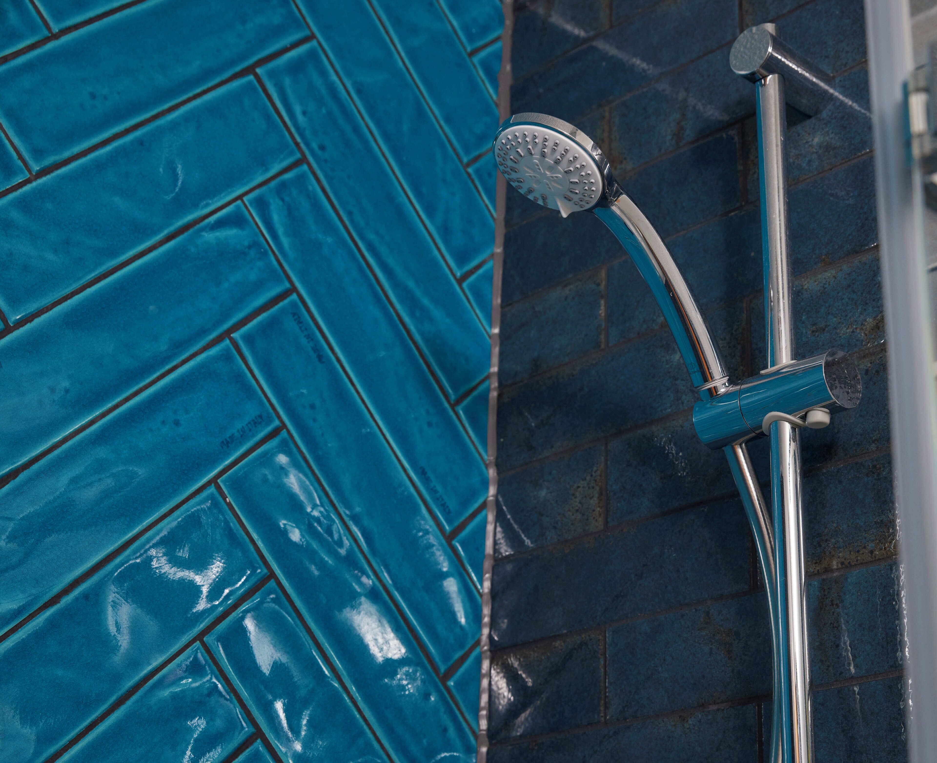 A shower with a chrome handle and a handheld showerhead, set against a blue tiled wall.