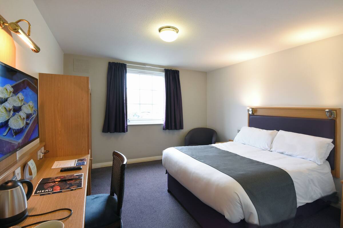 Una habitación de hotel con cama, escritorio, silla, televisión y ventana con cortinas.
