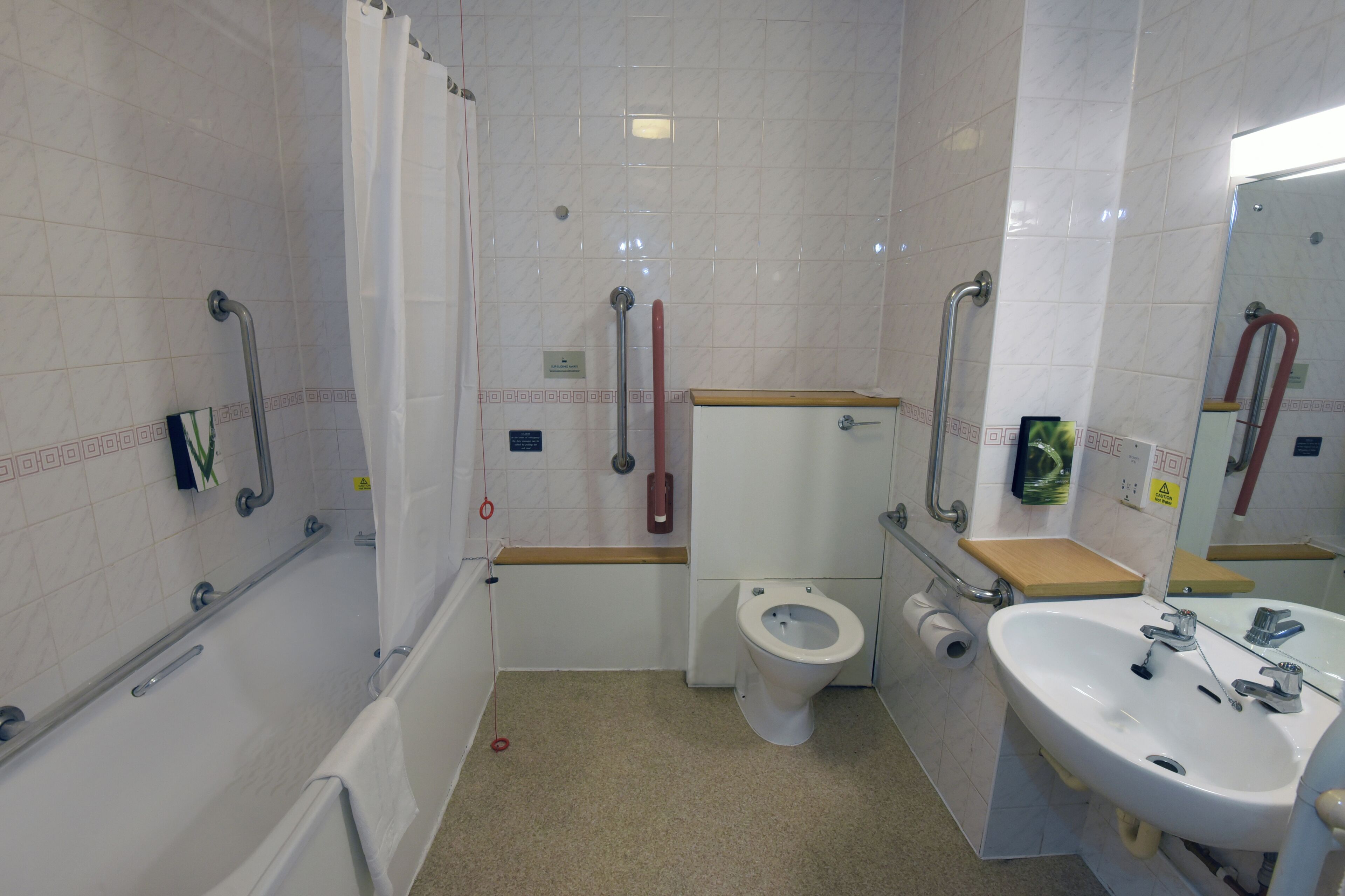 Ein Badezimmer mit Dusche, Toilette und Waschbecken, ausgestattet mit Haltegriffen und einer rollstuhlgerechten Dusche.
