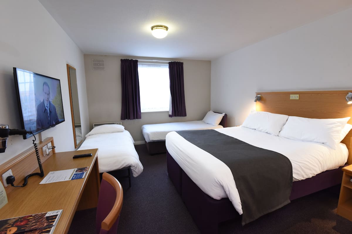 Una habitación de hotel con una cama grande, dos camas más pequeñas, un escritorio con silla, un televisor y una ventana con cortinas.