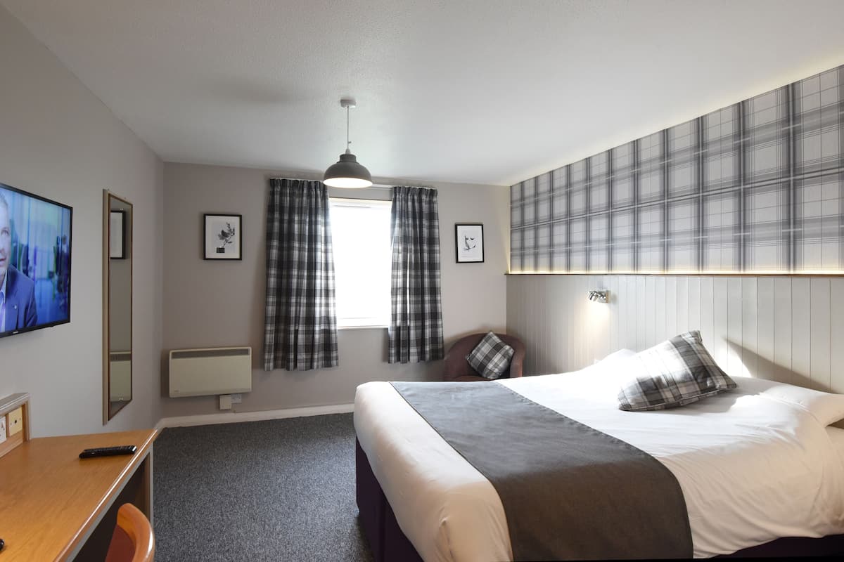 Habitación de hotel con cama, escritorio, silla, televisión, ventana con cortinas, lámpara de pared y radiador.