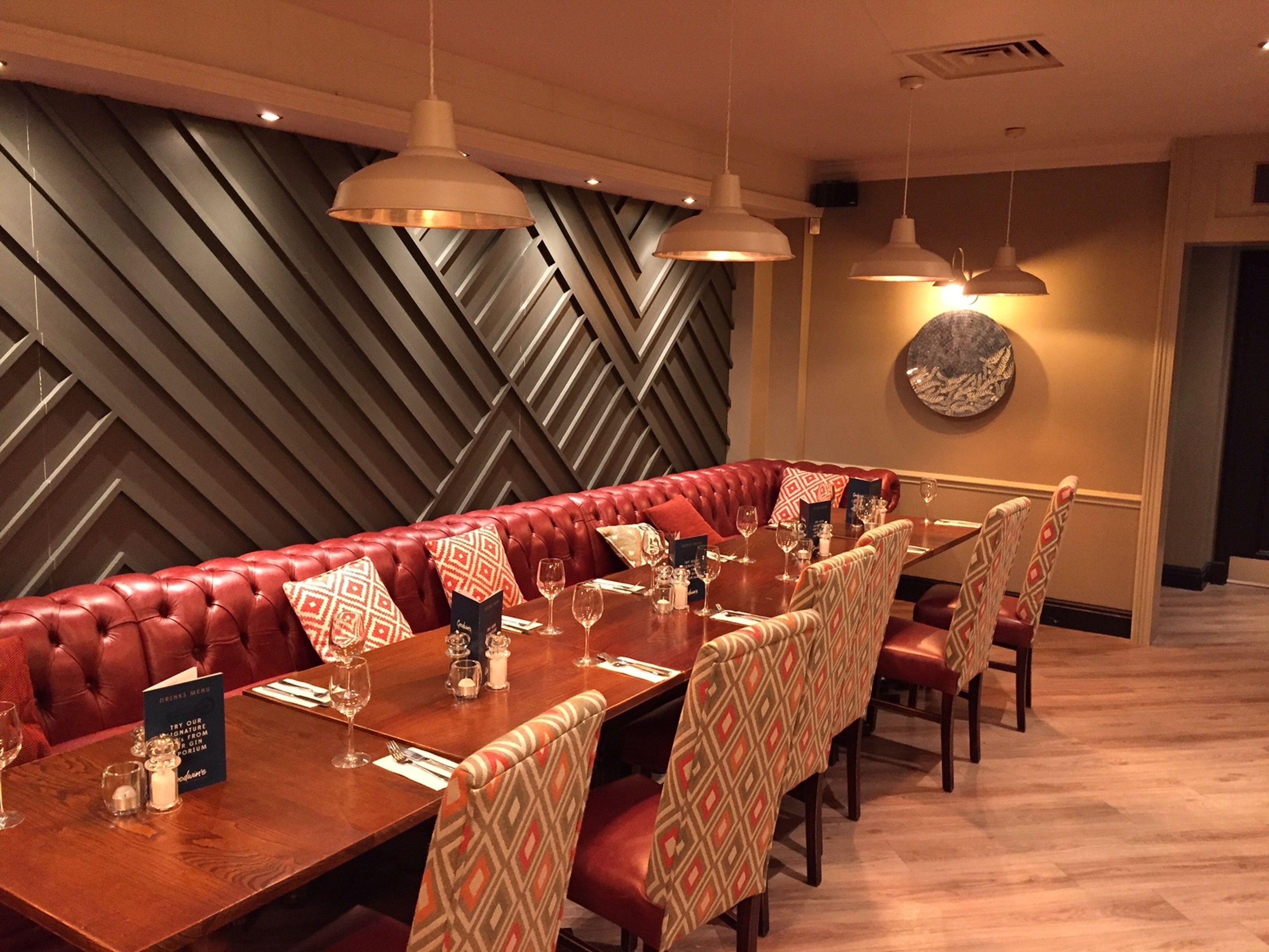 Un restaurante con una larga mesa de madera preparada para comer, asientos tapizados en rojo, sillas estampadas e iluminación colgante.