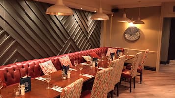 Un restaurante con una larga mesa de madera preparada para comer, asientos tapizados en rojo, sillas estampadas e iluminación colgante.
