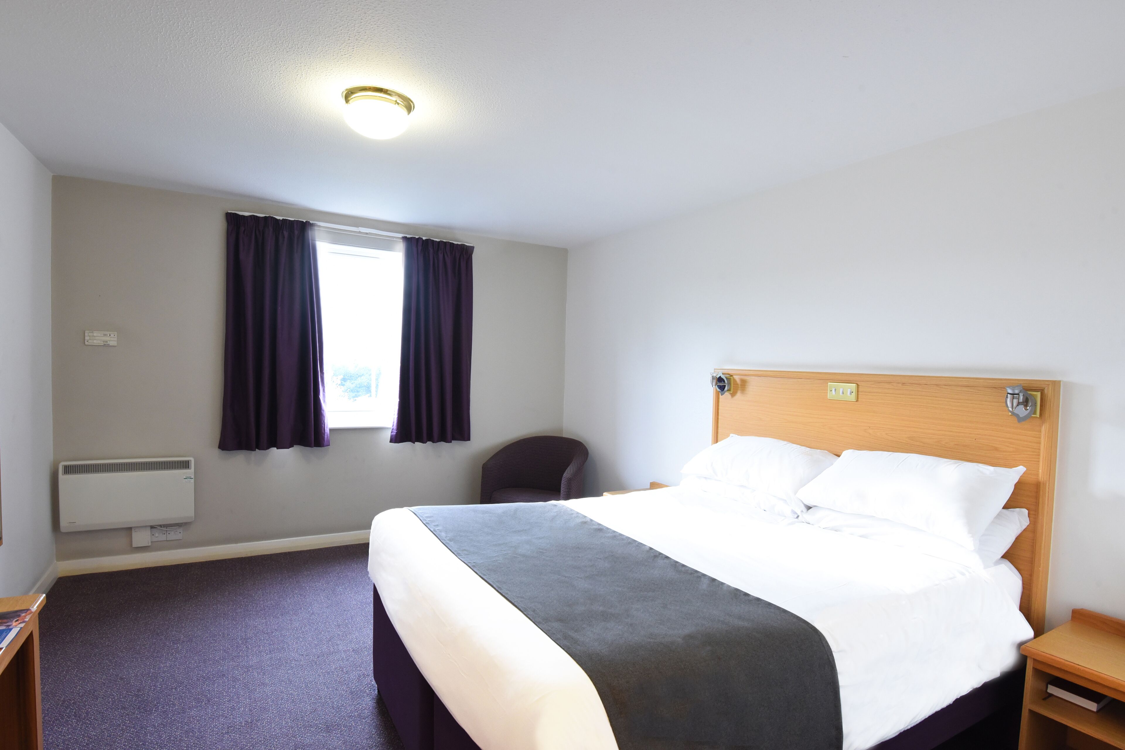 A hotel room with a bed, a chair, a nightstand, a window with curtains, and a ceiling light.
