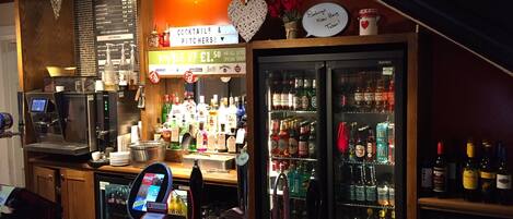A bar area with a coffee machine, a cash register, and a variety of bottles and glasses on shelves.