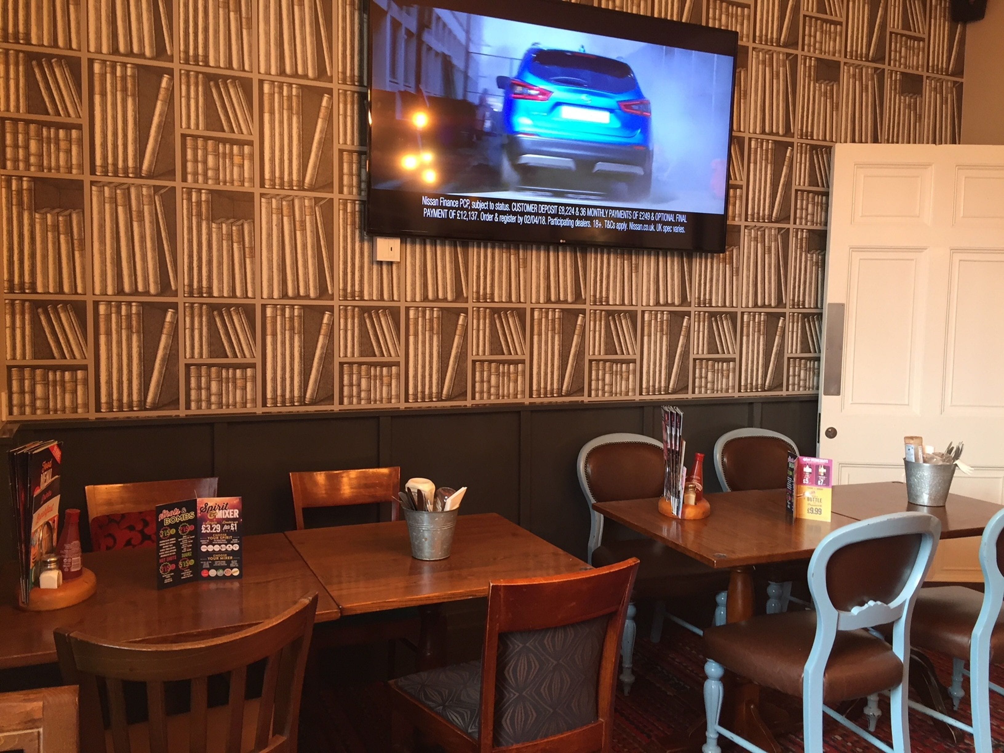 Interior de un restaurante con mesas y sillas de madera, un televisor en la pared mostrando un anuncio de automóvil, y una estantería llena de libros.