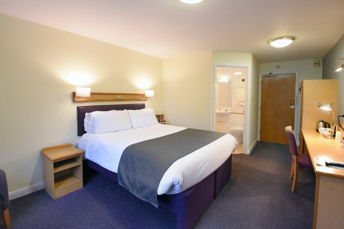 Ein Hotelzimmer mit Bett, Nachttisch, Lampe, Schreibtisch, Stuhl und einer Tür zu einem Badezimmer.