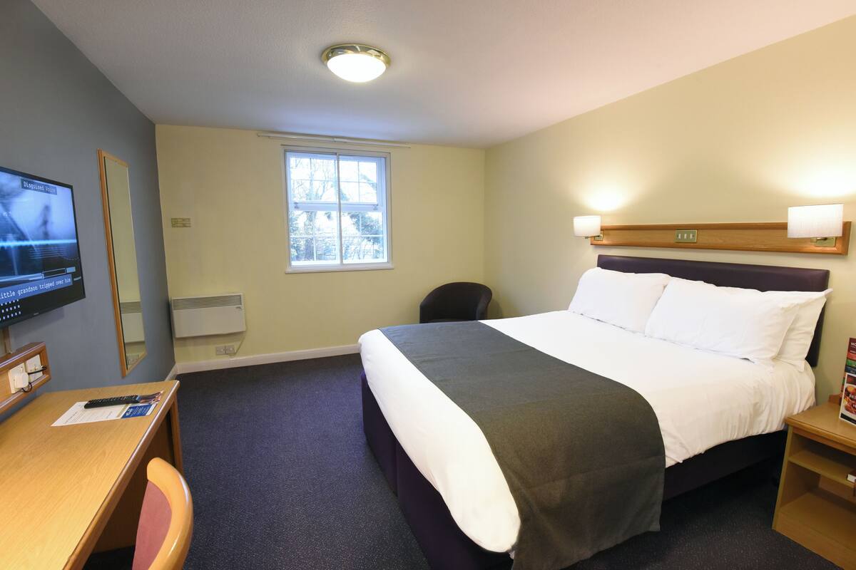 Ein Hotelzimmer mit Bett, Nachttisch, Schreibtisch, Stuhl, Fernseher, Fenster und Wandlampen.
