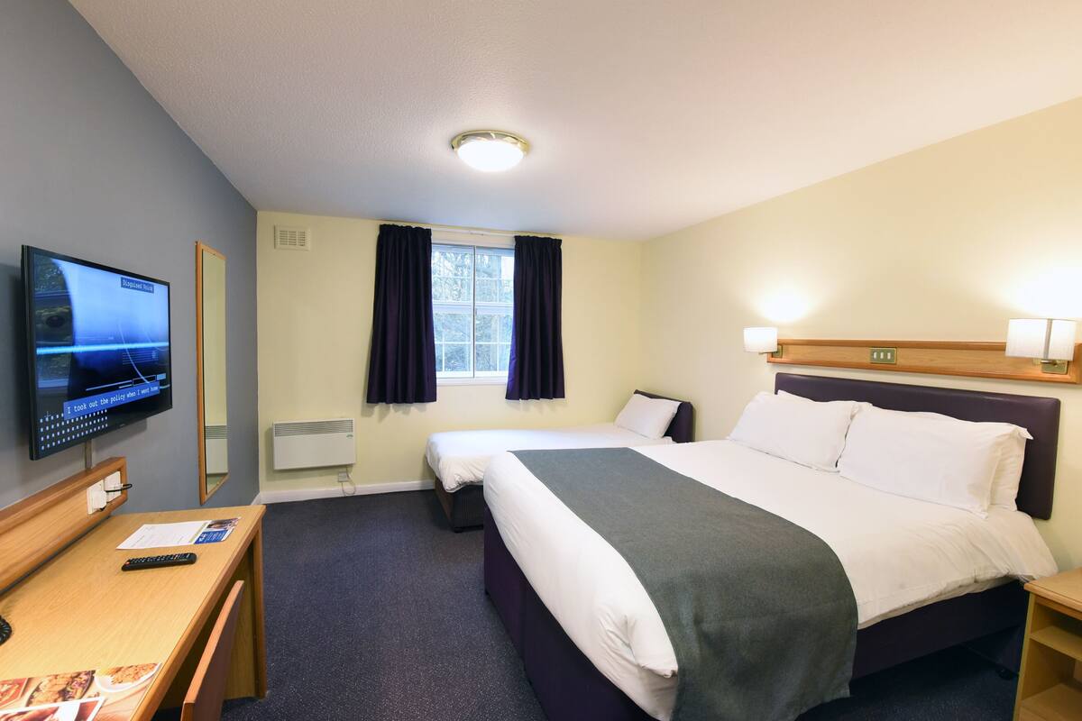 Ein Hotelzimmer mit einem Bett, einem Schreibtisch mit Computer, einem Fernseher, einem Fenster mit Vorhängen und einem Kopfteil mit Lampen.