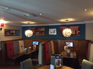 A restaurant with booths and tables, wooden paneling, hanging pendant lights, and wall-mounted televisions. - The Mount By Greene King Inns (Wigan)