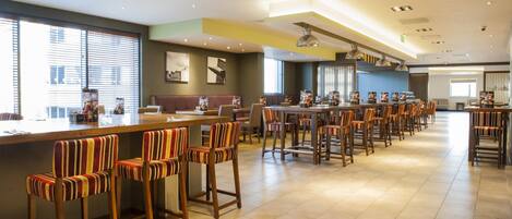A spacious hotel lobby with multiple seating areas, including bar stools and tables, and a large window providing natural light.