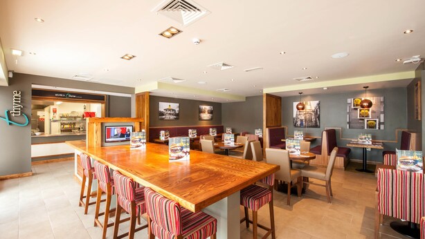 A restaurant with a long wooden bar, striped barstools, and a television screen. - Premier Inn Wigan Town Centre (Wigan)