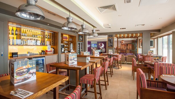 A restaurant with wooden tables and striped chairs, a bar area with bottles, and a menu board. - Premier Inn Wigan Town Centre (Wigan)