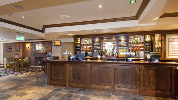 A bar area with a wooden counter, tiled flooring, and various bottles and glasses. - Premier Inn Glastonbury (Glastonbury)