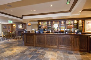 A bar area with a wooden counter, tiled flooring, and various bottles and glasses. - Premier Inn Glastonbury (Glastonbury)