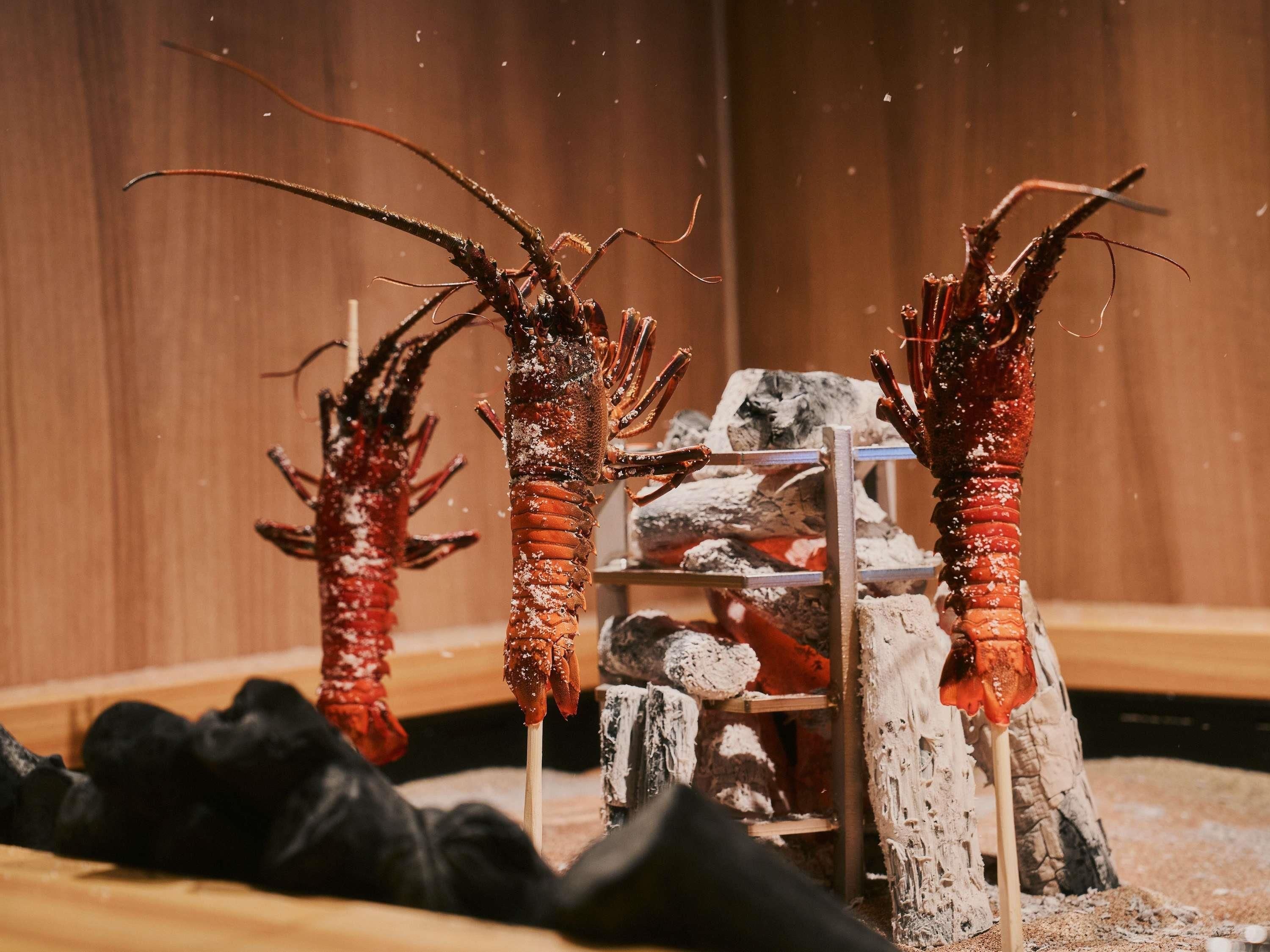 Two lobsters on skewers, with a wooden background and a shelf with charcoal.