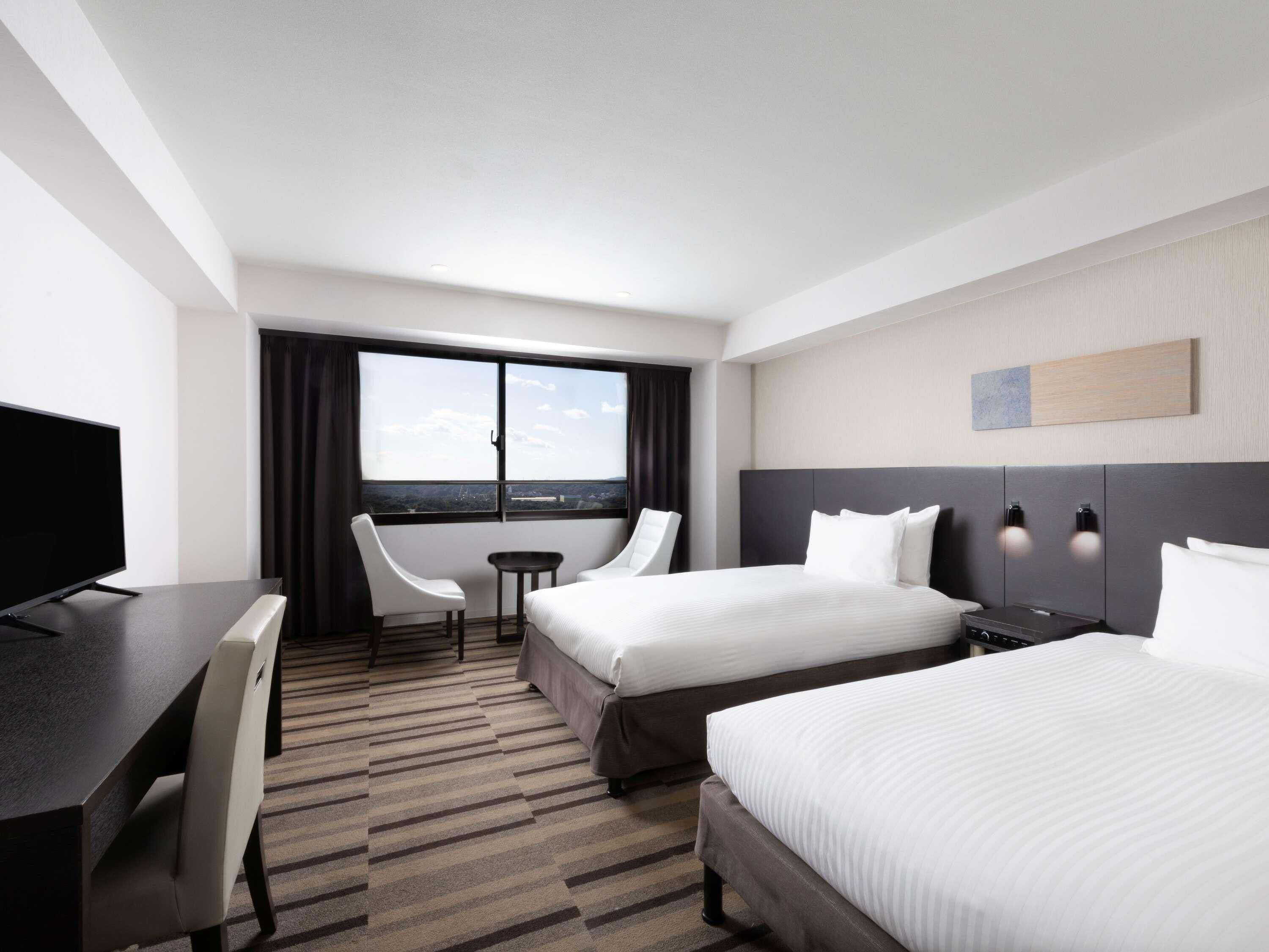 A hotel room with two beds, a desk, a chair, a TV, and a window with a view.