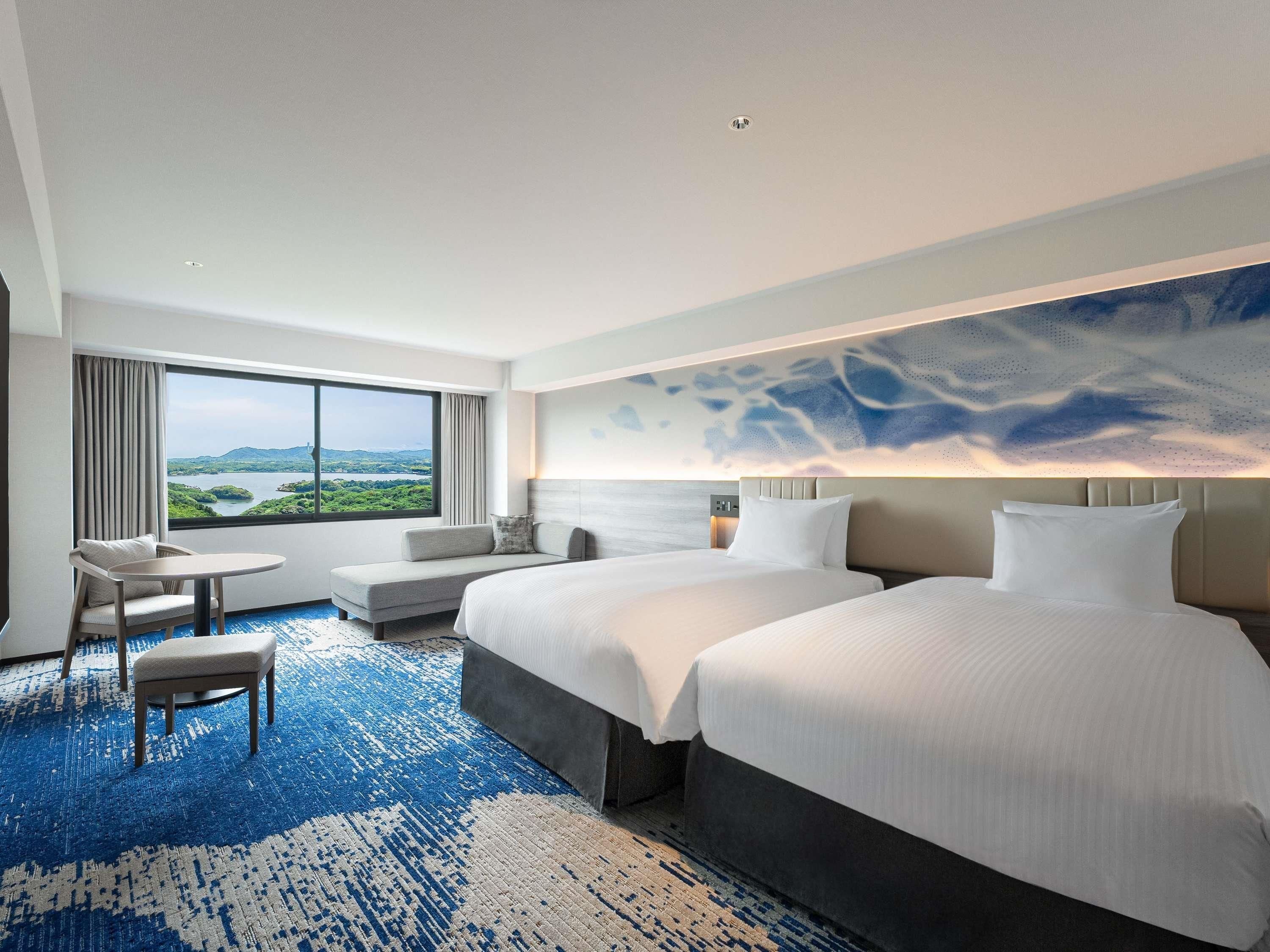 A hotel room with a large bed, a smaller bed, a sofa, a chair, and a table. There is a large window with a view of a landscape.