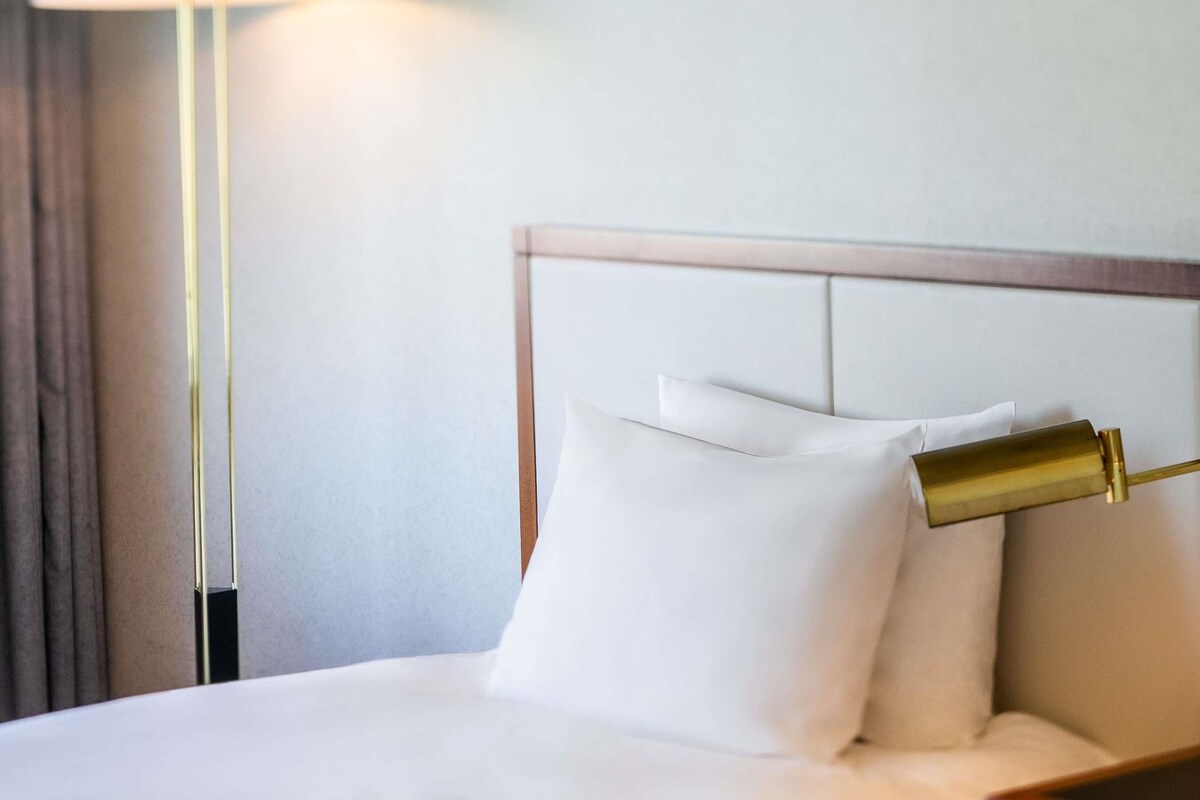 Una cama bien hecha con sábanas y almohadas blancas, cabecero dorado y una lámpara de pie con pantalla blanca.