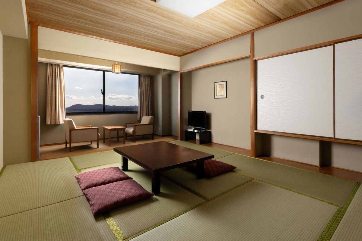 Una habitación japonesa tradicional con suelo de tatami, una mesa baja, un sofá, un televisor y una ventana con vistas.