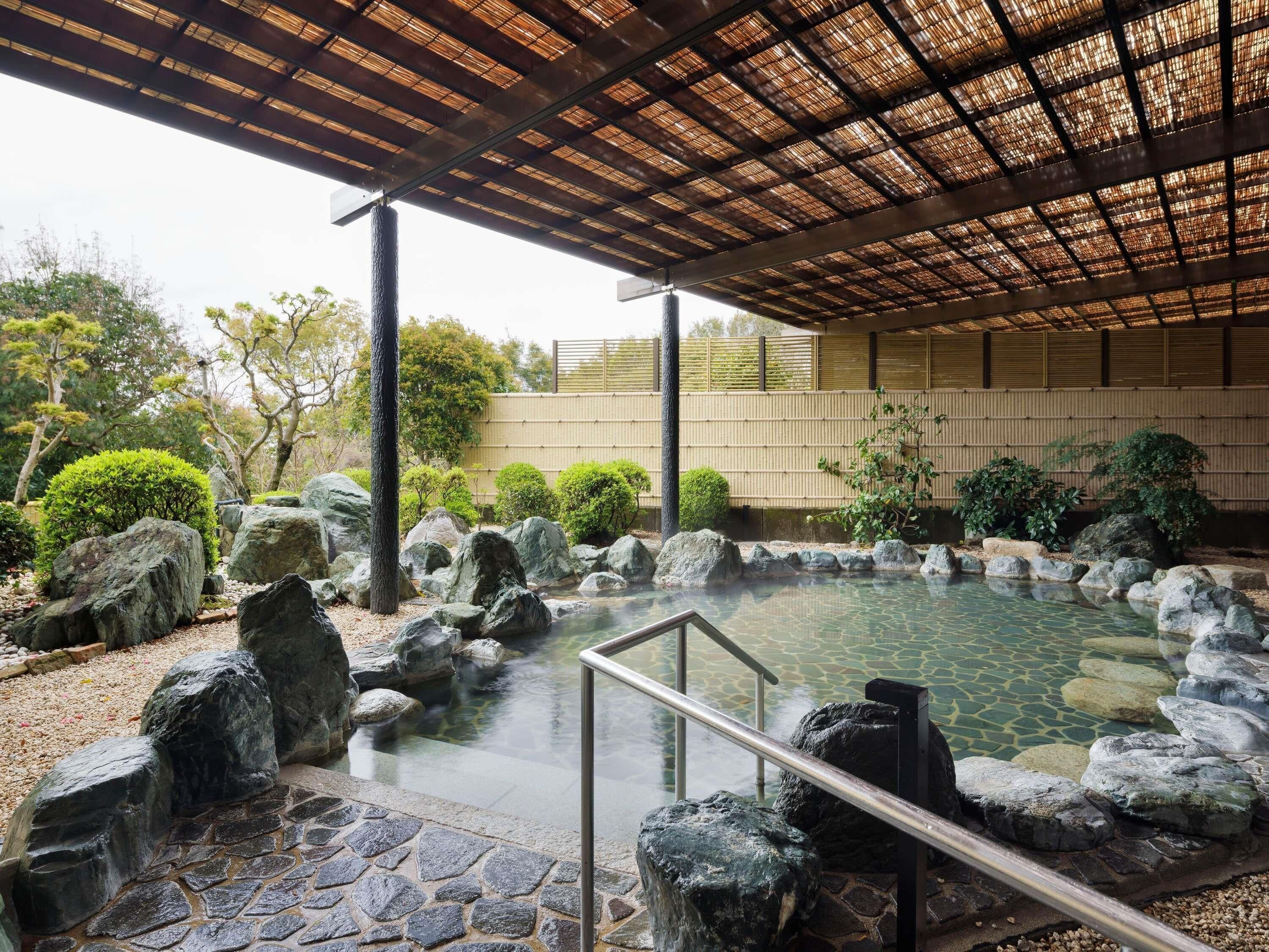 A traditional Japanese-style hot spring with a stone pathway, a wooden pergola, and surrounding greenery.