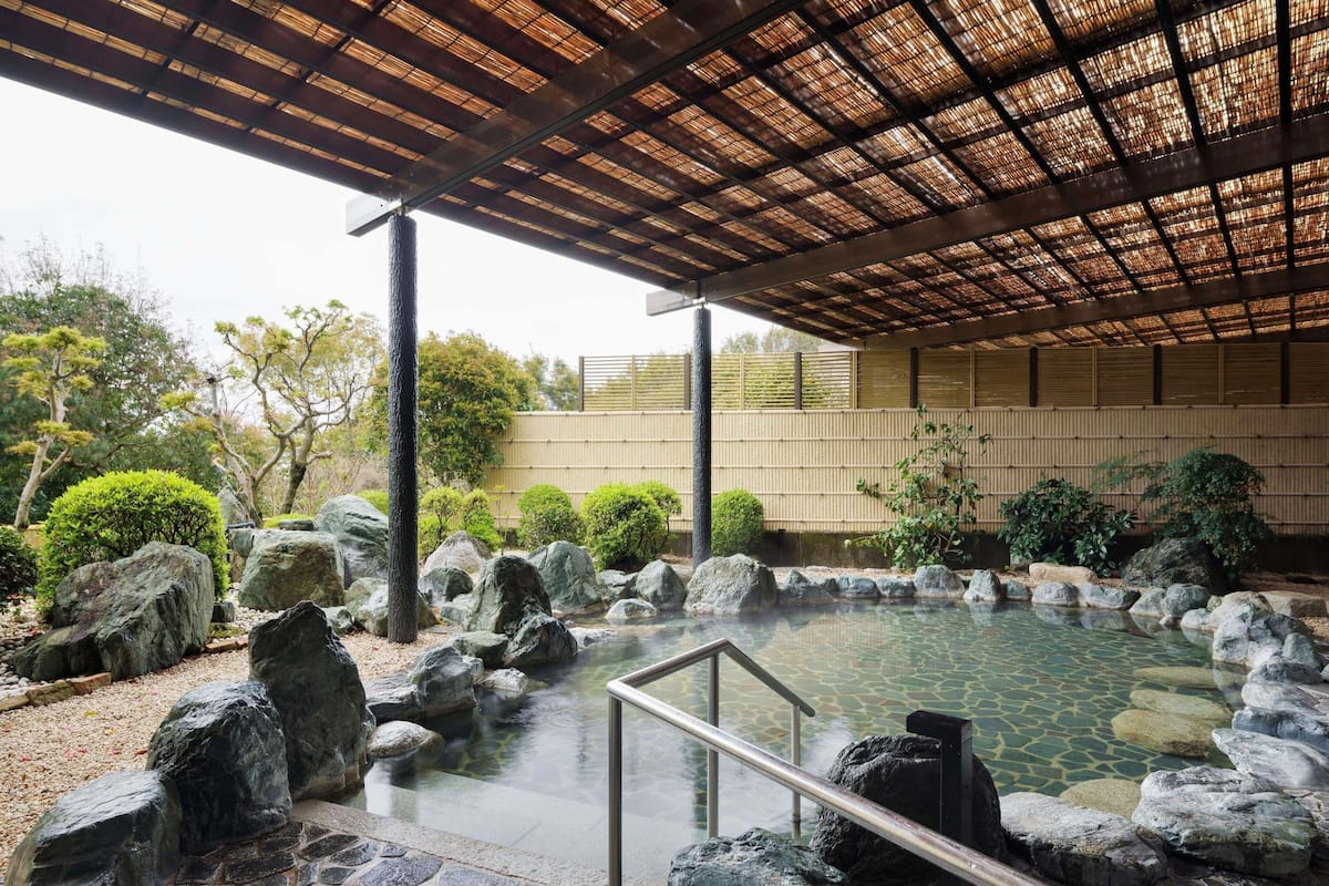 A traditional Japanese-style hot spring with a stone pathway, a wooden pergola, and surrounding greenery.