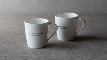 Two white mugs with 'Grand Mercure Hotels and Resorts' printed on them, placed on a textured surface.