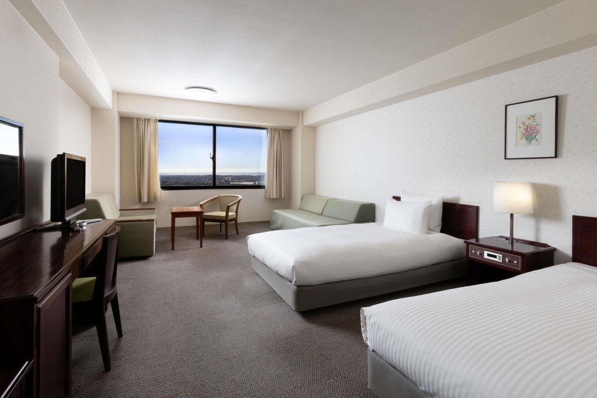 Habitación de hotel con cama, escritorio con televisor, silla, sofá y un amplio ventanal con vista a la ciudad.