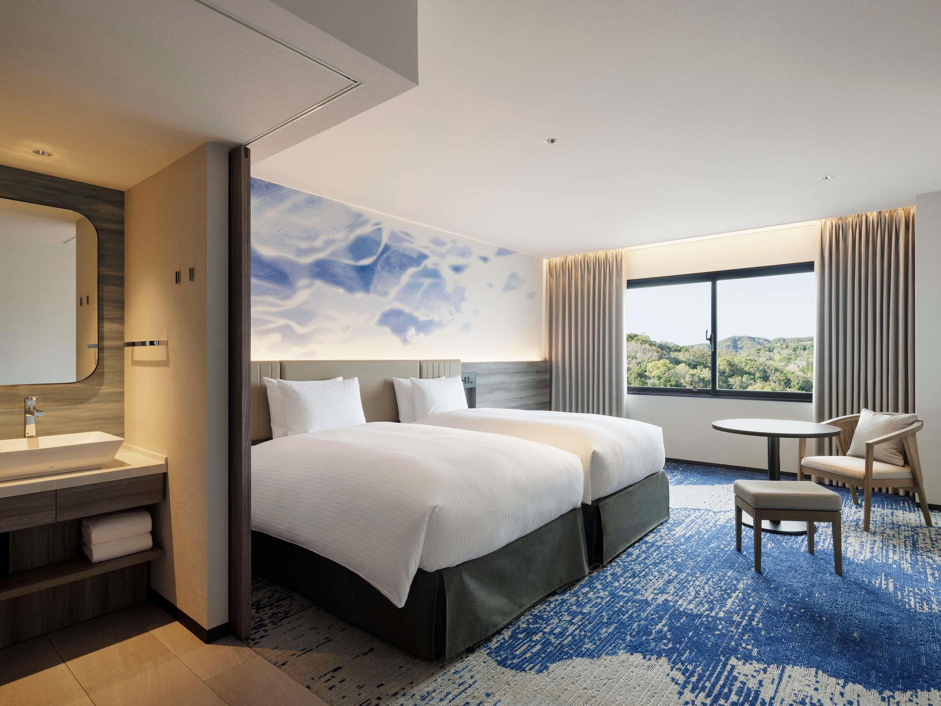 Hotelkamer met een groot bed, nachtkastje, stoel, klein tafeltje, badkamer met wastafel en spiegel, en een raam met uitzicht op groen.