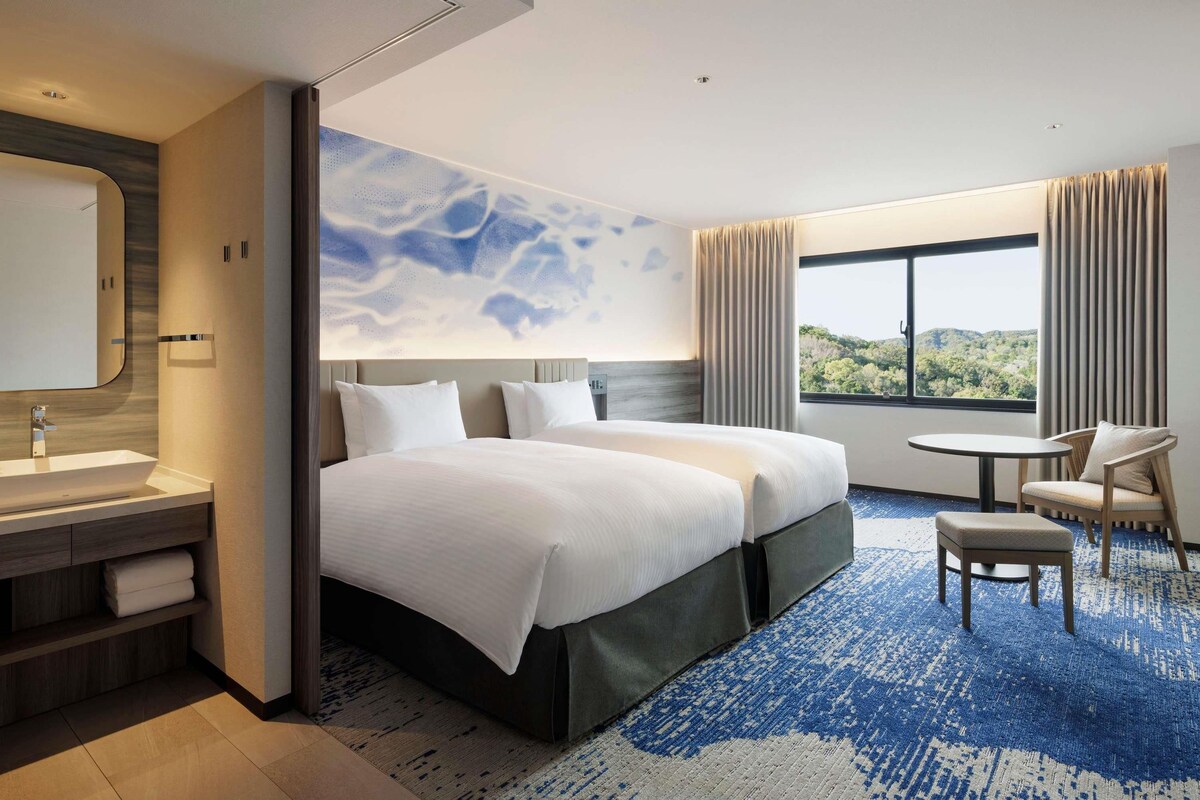 Habitación de hotel con una cama grande, una mesita de noche, una silla, una pequeña mesa, un baño con lavamanos y espejo, y una ventana con vistas a zonas verdes.