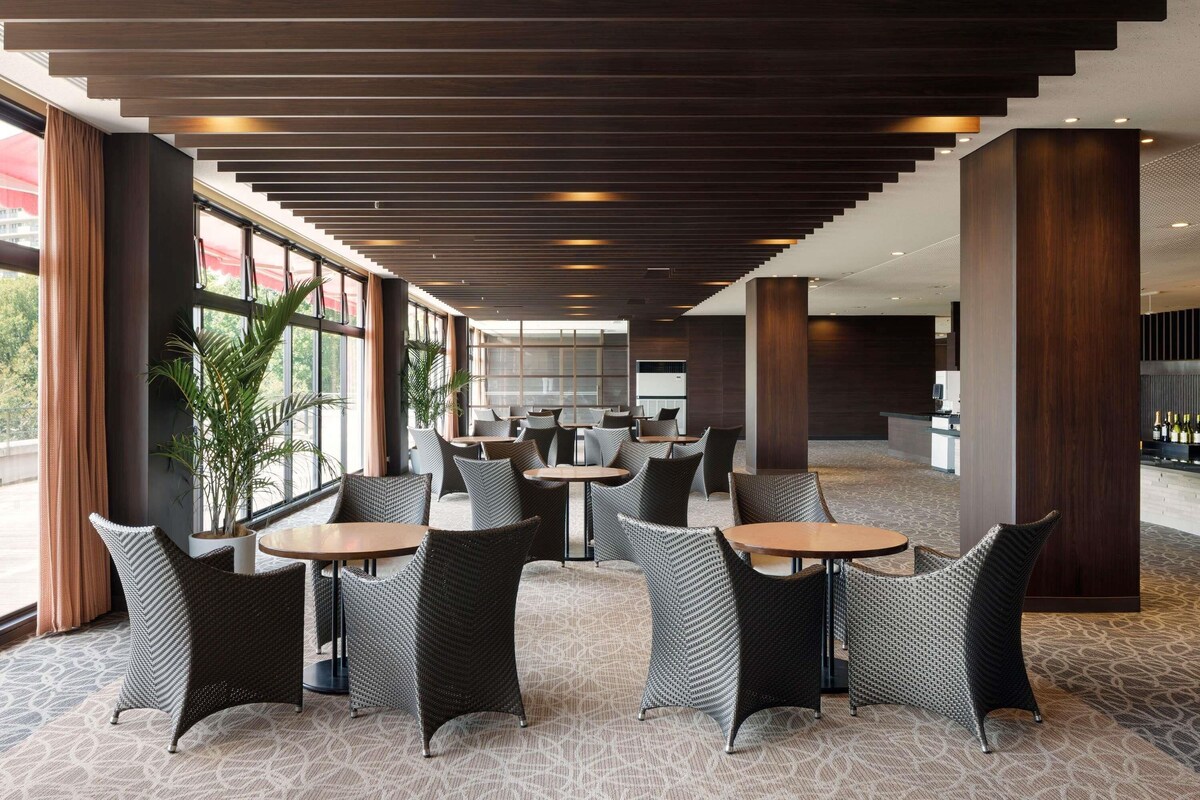 Un hall d'hôtel moderne, doté de meubles en osier, de grandes fenêtres et d'un tapis à motifs.