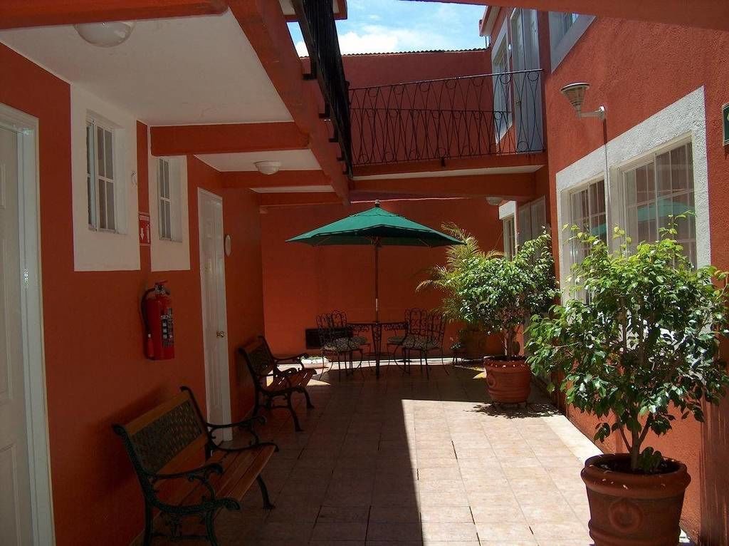 A courtyard with a bench, chairs, a table, and a green umbrella. There are potted plants and a fire extinguisher on the wall.