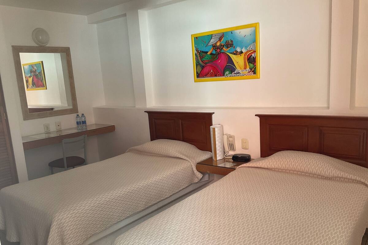 A hotel room with two beds, a bedside table with a phone and a lamp, a mirror, and a wall with a colorful painting.