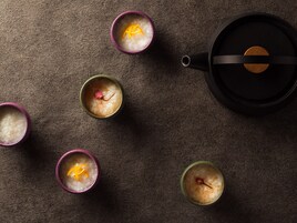 A top-down view of a table with five cups of a clear liquid, possibly a beverage, each with a slice of orange and a red flower. A black teapot is placed to the right.