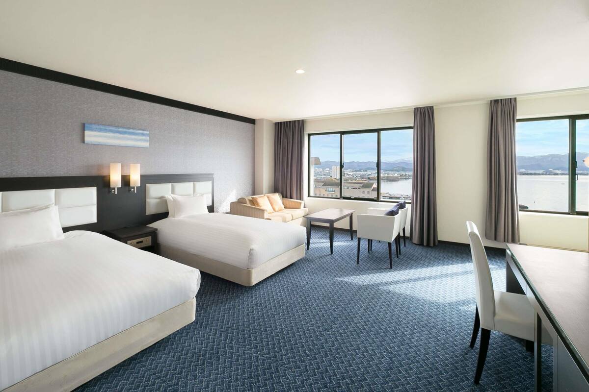 Habitación de hotel con dos camas, un sofá, una mesa de comedor y vistas a la ciudad a través de amplias ventanas.