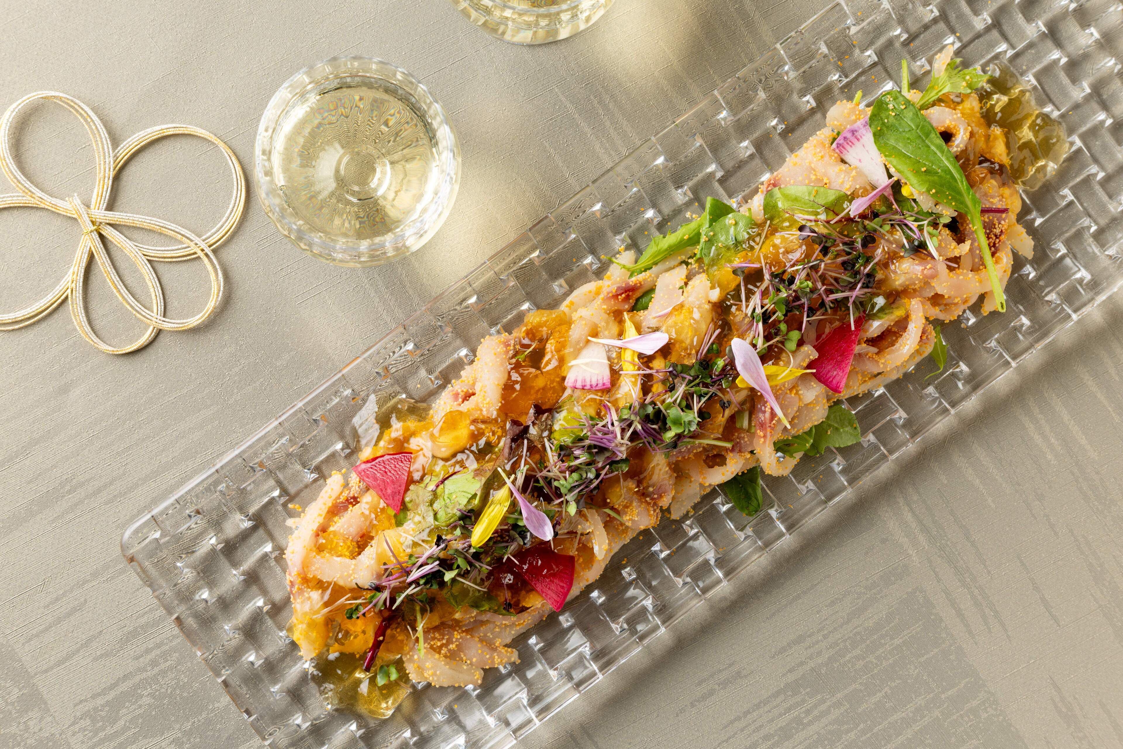 A glass plate with a seafood dish garnished with greens and pink flowers, accompanied by a glass of white wine and a gold decorative item.