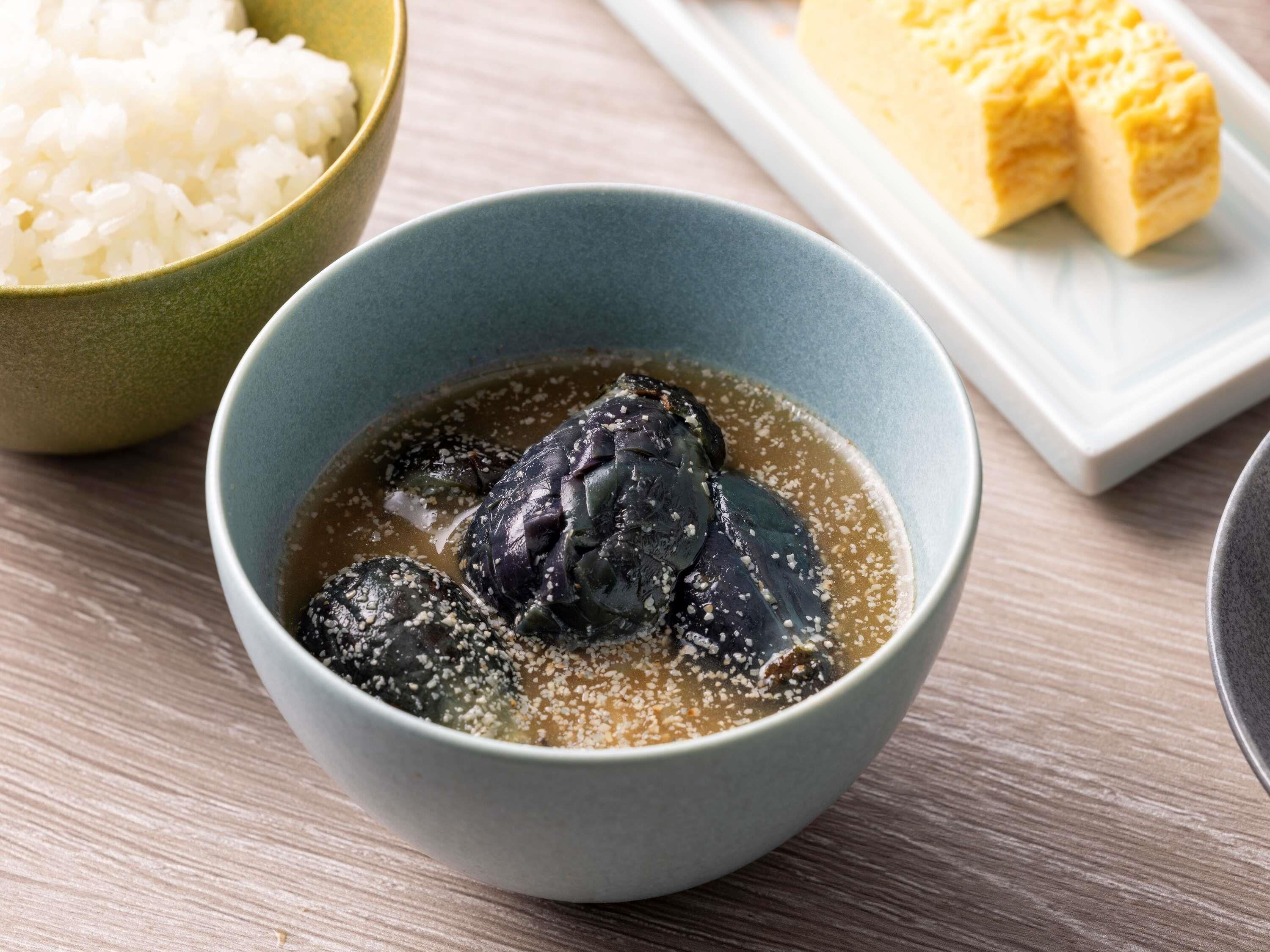 A bowl of dark-colored soup with a garnish, a bowl of white rice, and a block of yellowish food on a white plate.