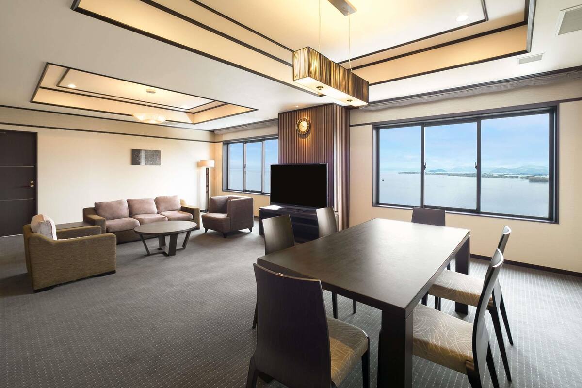 Habitación de hotel moderna con un ventanal grande, televisor de pantalla plana, zona de comedor y rincón de estar con sofás y sillones.